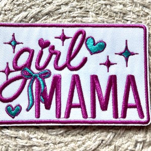 May include: Embroidered patch with the words "girl MAMA" in a cursive and block font. The patch is white with a pink border and features pink text, teal hearts, stars, and a bow.