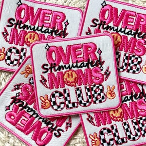 May include: Several square patches with the text "OVER Stimulated MOMS CLUB" in pink, black, and white. The patches feature a smiley face, peace signs, and a checkered pattern. Each patch has a pink border.