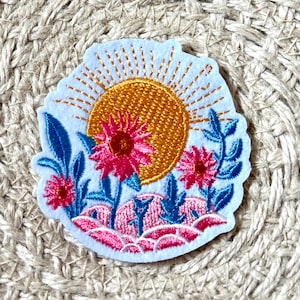 Puede incluir: Parche bordado con un sol dorado, flores rosas y hojas azules. El parche tiene un borde azul claro y está sobre un fondo tejido neutro. El diseño evoca un tema de naturaleza o floral. Aproximadamente 7 cm de diámetro.