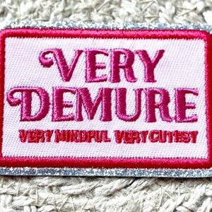 May include: Embroidered patch with the words "VERY DEMURE" in pink script. The patch has a pink background, a red border, and a silver glitter edge. The text "VERY MINDFUL VERY CUTEST" is below the main text.