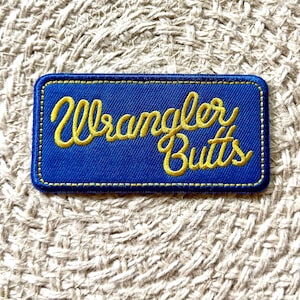 May include: A rectangular embroidered patch with a royal blue background and a yellow stitched border. The text Wrangler Butts is embroidered in a yellow cursive script across the center of the patch, featuring a clean, finished edge design.