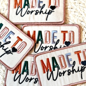 以下が含まれることがあります： 「MADE TO Worship」という文字と小さなハートが書かれた、カラフルな布のパッチが4つ。パッチはベージュの布でできており、縁はピンクのステッチで縫われています。