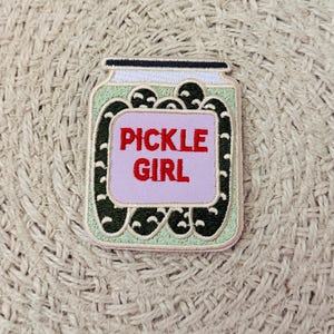 May include: An embroidered patch shaped like a glass jar filled with dark green pickles. The jar features a light purple rectangular label in the center with the red text PICKLE GIRL printed on it. The patch has a light green background and a dark blue lid.