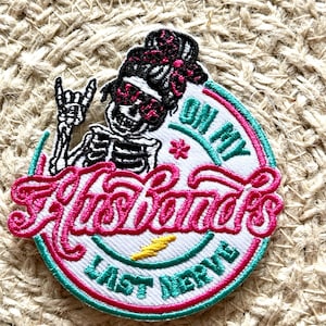 May include: A white embroidered patch with a pink skeleton wearing sunglasses and a pink bandana. The text "On My Last Nerve" is written in pink around the skeleton. A yellow lightning bolt is below the text.