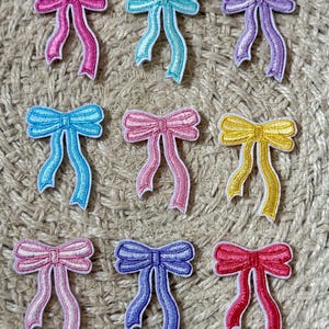 May include: Nine embroidered bow patches in various colors, including pink, blue, purple, and yellow. Each bow has a white outline and flowing ribbons. The patches are arranged on a textured, neutral-toned surface.