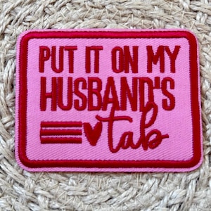 Può includere: Toppa rettangolare rosa con bordo rosso e testo ricamato. Il testo recita "PUT IT ON MY HUSBAND'S tab" in rosso. La toppa ha un cuore e tre linee orizzontali.