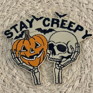 May include: An embroidered patch featuring a beige background with the black text STAY CREEPY and black silhouettes of bats. The design includes an orange jack-o'-lantern pumpkin held by a skeleton hand and a beige human skull.