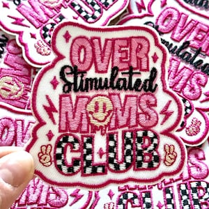 May include: Pink and black embroidered patch with the text "Over Stimulated Moms Club" in a white cloud shape. The text is in a bold, cursive font. The patch has a checkered pattern and a peace sign.