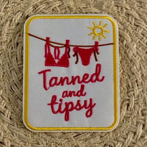 Puede incluir: Un parche bordado rectangular con fondo blanco y borde amarillo. Presenta un bikini rojo colgado en un tendedero, un sol amarillo y el texto bordado en rojo Tanned and tipsy en una fuente de estilo cursiva.