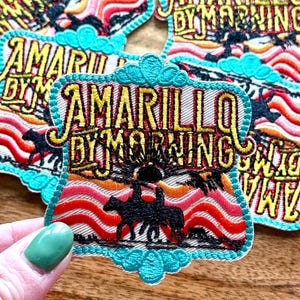 May include: Embroidered patch with the text "Amarillo By Morning" in yellow and black. The patch features a sunset scene with two riders on horseback. The patch has a turquoise border and is on a wooden surface.