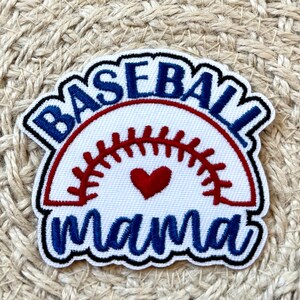 May include: A baseball-themed patch with the words "BASEBALL MAMA" in blue, outlined in black. The design includes a red baseball stitch and a red heart. The patch is on a textured, beige background.