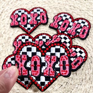 May include: A set of six pink and black heart-shaped patches with the word "XOXO" embroidered in pink on a black and white chequered background.