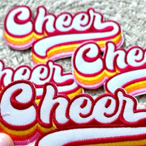 May include: Embroidered patches with the word "Cheer" in a retro script font. The patches feature a white background with red trim and a gradient of yellow, orange, and pink stripes.