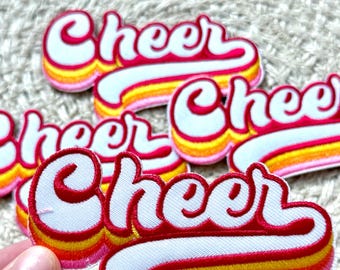 May include: Embroidered patches with the word "Cheer" in a retro script font. The patches feature a white background with red trim and a gradient of yellow, orange, and pink stripes.