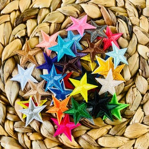 May include: A collection of embroidered star patches in various colors, including pink, blue, yellow, and green. The patches are arranged on a woven, textured background, creating a vibrant and eye-catching display. These patches are ideal for adding a decorative touch to clothing or accessories.