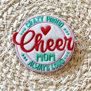 Puede incluir: Un parche bordado redondo con las palabras "Crazy Proud Cheer Mom Always Loved". La palabra "Cheer" está en escritura roja grande, con un corazón rojo encima. El parche tiene un fondo blanco con un borde rosa y verde.