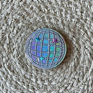 Può includere: Una toppa a forma di palla da discoteca argentata con glitter e tre stelle iridescenti. La toppa è su uno sfondo di tessuto intrecciato.