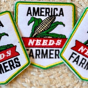 May include: Three embroidered patches with a white background, green and yellow border, and the text "AMERICA NEEDS FARMERS". Each patch features a corn graphic with green leaves and a red banner.