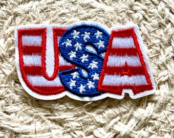 Può includere: Toppa ricamata con le lettere "USA" in rosso, bianco e blu. Le lettere "U" e "A" hanno strisce rosse e bianche, mentre la "S" è blu con stelle bianche. La toppa è su uno sfondo beige strutturato.