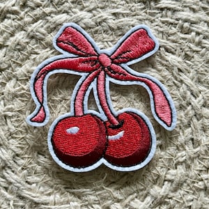 Può includere: Una toppa ricamata di ciliegie rosse con un fiocco rosa. Le ciliegie sono ricamate con un effetto 3D e il fiocco è piatto.