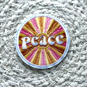 Puede incluir: Parche bordado circular con la palabra "Peace" en escritura blanca. El parche presenta un diseño de rayos de sol en tonos rosa, dorado y marrón, con un borde blanco. Ideal para ropa o accesorios.
