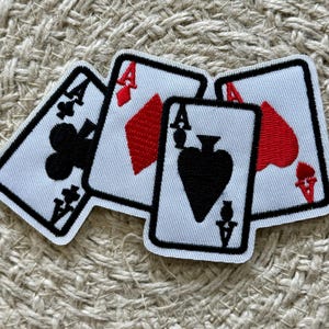 Puede incluir: Cuatro parches bordados para planchar de cartas de juego, cada uno con un palo diferente: tréboles, diamantes, corazones y picas. Cada parche tiene un fondo blanco con contornos negros y símbolos rojos.