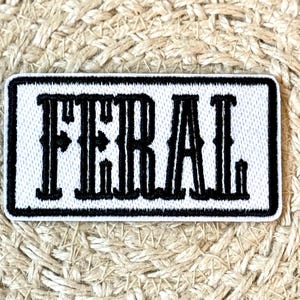 May include: Embroidered patch with the word "FERAL" in bold, black, stylized letters on a white background. The patch has a black border and is rectangular. The background is a woven, beige material.