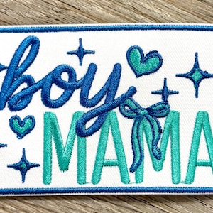 Puede incluir: Parche bordado con las palabras "boy MAMA" en azul y verde azulado. El parche presenta corazones, estrellas y un diseño de lazo. El parche tiene un fondo blanco con un borde azul.