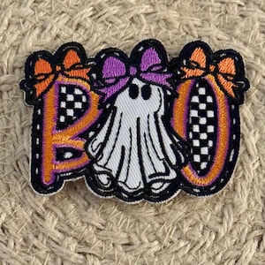 May include: An embroidered patch featuring the word BOO in orange and black with a white ghost in the center. The letters B and O are decorated with orange bows, while the ghost wears a purple bow. The design includes a black and white checkerboard pattern.