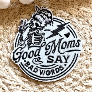 Puede incluir: Parche blanco bordado con un esqueleto negro sosteniendo un signo de paz. El texto dice "Good Moms Say Bad Words" con un rayo y un corazón.