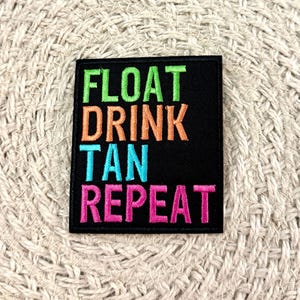 Puede incluir: Un parche rectangular negro bordado con el texto FLOAT en verde lima, DRINK en naranja, TAN en cian y REPEAT en rosa intenso. Las letras están apiladas en cuatro líneas, con cada palabra representada en un estilo de fuente sans-serif en negrita.