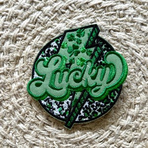 May include: Embroidered patch with a green and black design. The patch features the word "Lucky" in a stylized font, a green lightning bolt, and a background with a leopard print pattern. The patch is circular and has a black border.