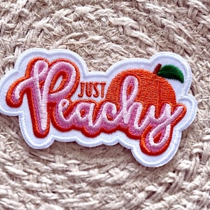 Könnte beinhalten: Gestickter Aufnäher mit den Worten "Just Peachy" in rosa und roter Schrift, mit einer orangefarbenen Pfirsich mit einem grünen Blatt. Der Aufnäher hat einen weißen Rand und befindet sich auf einer strukturierten beigefarbenen Oberfläche.