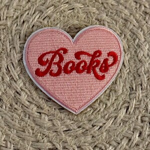 May include: A heart-shaped embroidered patch featuring a light pink background with a white border. The word Books is stitched across the center in a bold, red, cursive script font. The patch has a textured surface and a distinct fabric edge.