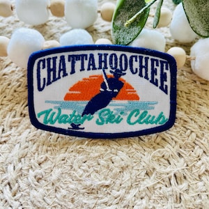 May include: A white embroidered patch with blue and green accents. The patch features a silhouette of a person water skiing with a sunset in the background. The text "Chattahoochee Water Ski Club" is embroidered in blue.