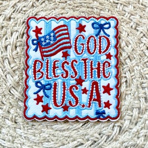 Puede incluir: Un parche cuadrado con borde festoneado, con el texto "GOD BLESS THE USA" en rojo, blanco y azul. El diseño incluye una bandera americana, estrellas y lazos, todo sobre un fondo azul a rayas.
