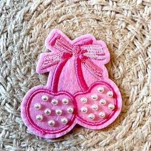 May include: A pink felt cherry patch with a bow and pearl accents. The cherries are embellished with small white pearls and outlined with a darker pink thread. The bow is also pink and has detailed stitching.
