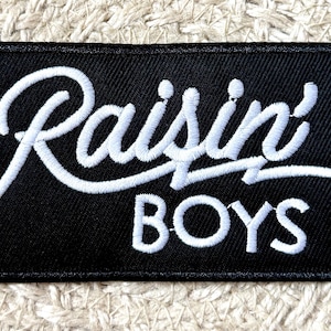 Könnte beinhalten: Schwarzer rechteckiger Aufnäher mit den Worten "Raisin' BOYS" in weißer Schrift gestickt. Der Aufnäher hat einen schwarzen Rand und befindet sich auf einem strukturierten, cremefarbenen Hintergrund.
