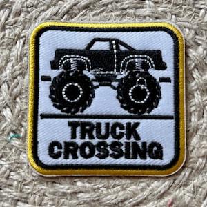 May include: Square patch with a black monster truck design and the words "TRUCK CROSSING" in black text. The patch has a white background with a yellow border.
