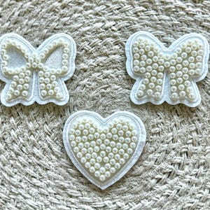 Könnte beinhalten: Drei weiße Stoff-Patches mit Perlenbesatz. Die Patches sind herzförmig, schleifenförmig und schleifenförmig.