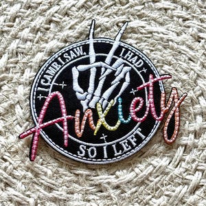 Pode incluir: Um remendo bordado preto e branco com uma mão de esqueleto fazendo um sinal de paz. O texto ao redor da mão diz "Eu vim, eu vi, eu tive ansiedade, então eu fui embora". O texto "ansiedade" está em cores do arco-íris.