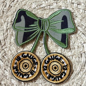 May include: Embroidered patch featuring a green bow and two cherry-like circles. The bow has a camouflage pattern. The circles have a gold center with the text "12 GAUGE" in a circular pattern.