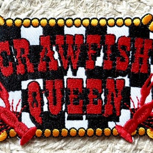 May include: Embroidered patch with the words "CRAWFISH QUEEN" in red, outlined in black, on a black and white checkered background. The patch features red crawfish and a yellow and orange border.