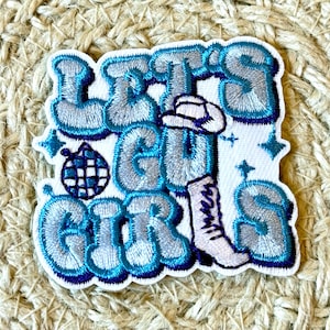 Puede incluir: Parche blanco para planchar con bordado azul. El parche presenta el texto "Let's Go Girls" en una fuente estilizada con una bola de discoteca y una bota con un sombrero de vaquero.