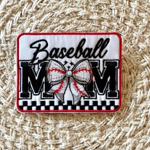Könnte beinhalten: Gestickter Aufnäher mit den Worten "Baseball Mom" in schwarzer Schrift und Blockbuchstaben. In der Mitte befindet sich eine Baseball-Schleife mit roten Nahtdetails. Der Aufnäher hat einen rot-schwarzen Rand.