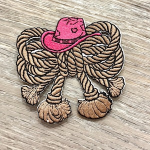 May include: Embroidered patch featuring a pink cowboy hat atop a tan rope design. The hat has a detailed brim and crown, with black outlining the hat and rope. The rope is intricately knotted, with detailed tassels at the bottom.