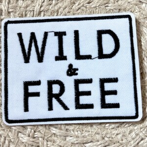 May include: A white rectangular patch with a black border. The patch features the words "WILD & FREE" in bold, black capital letters. The patch is likely an iron-on or sew-on patch for clothing or accessories.