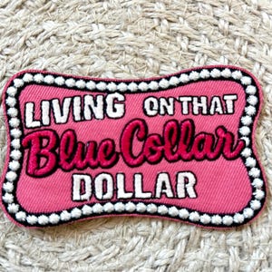 Può includere: Una toppa rosa con le parole "LIVING ON THAT Blue Collar DOLLAR" in lettere bianche e rosa acceso. La toppa ha un bordo di perline bianche ed è posta su uno sfondo strutturato.