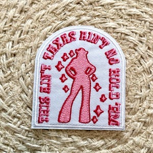 Peut inclure: Patch thermocollant blanc avec un texte brodé rose qui dit "This ain't Texas, hun, so hold your horses" et une silhouette rose d'une femme avec des étoiles autour d'elle.