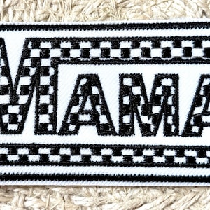 May include: A rectangular patch with the word "MAMA" in bold, black letters. The letters are filled with a checkered pattern. The patch has a white background and a black border with a checkered design.